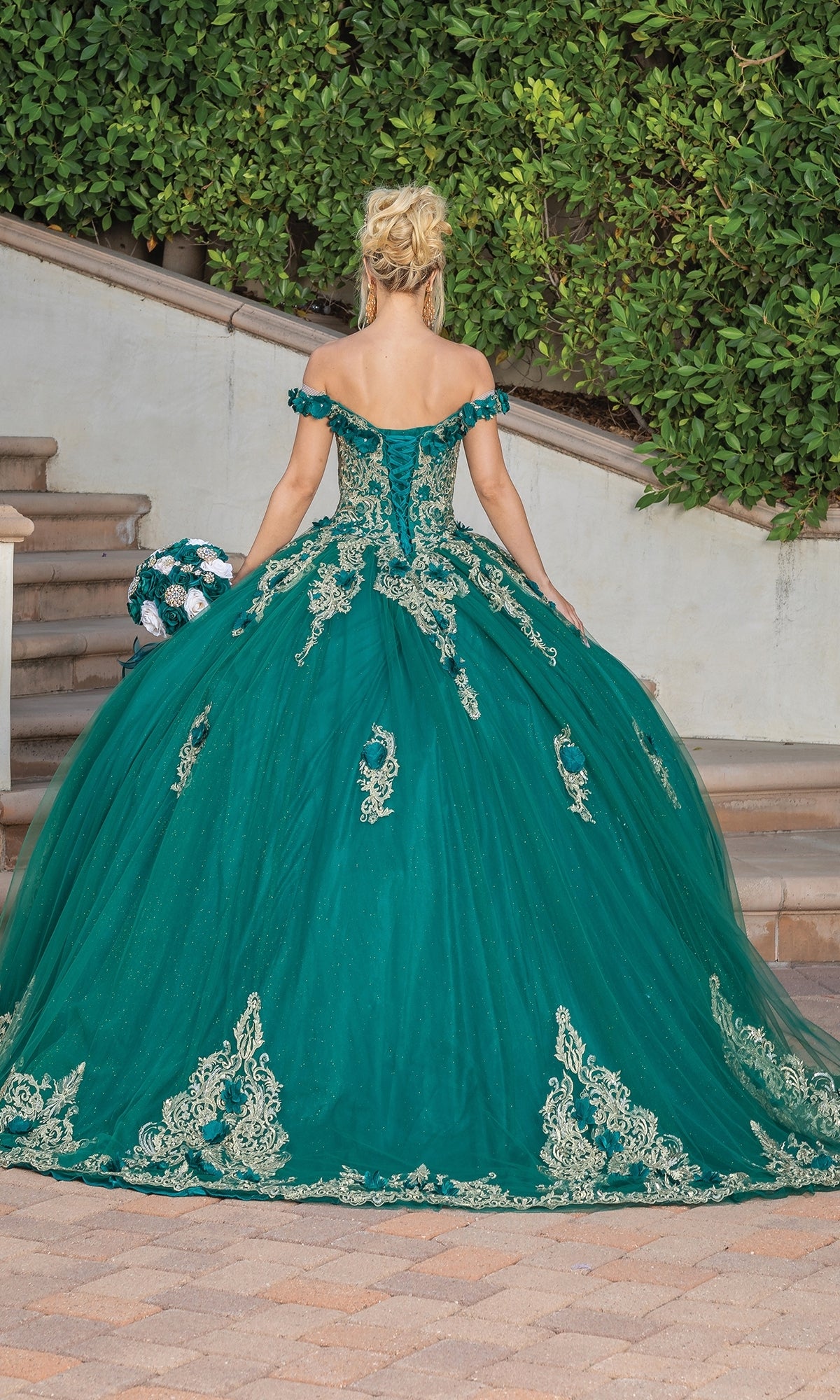 A woman stands outdoors, facing away, wearing the Quinceanera Dress 1739 by Dancing Queen—an off-the-shoulder gown with gold lace embroidery—and holding a matching bouquet.
